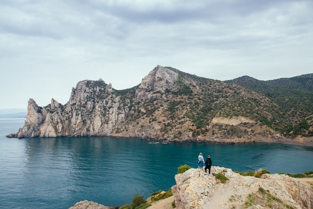 two-person-standing-on-rocky-hill-viewing-blue-sea